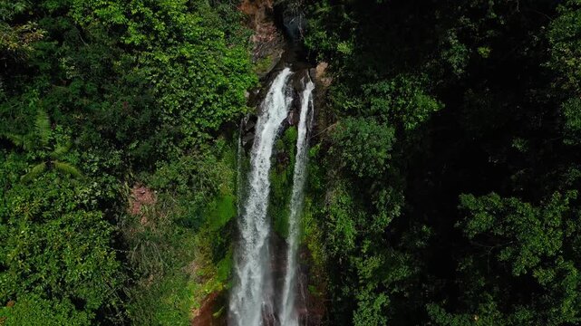 Closer drone footage of Gitgit Waterfall in Bali, fully visible and surrounded by dense green tropical trees and foliage, with clear blue sky and clouds adding to the stunning natural view.