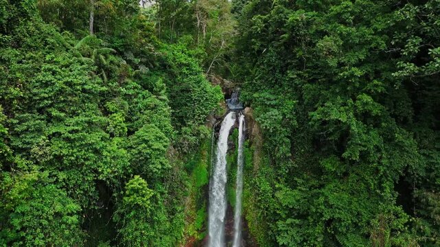Drone footage of Gitgit Waterfall in Bali, captured from a closer angle, showcasing powerful tropical water flow surrounded by dense green trees and foliage in a vibrant natural setting.