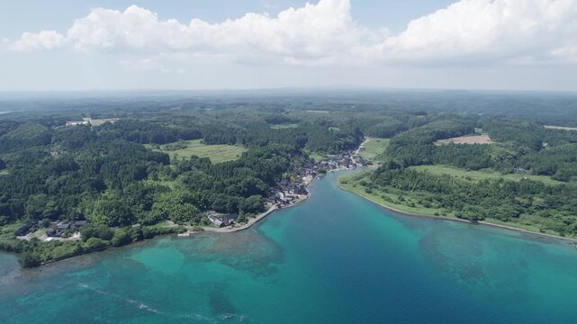 Drone footage of Anamizu, Sora area in Noto, Japan. The image captures a quiet coastal village, lush green forest, and turquoise sea blending into scenic inland farmland. Peaceful summer atmosphere.
