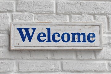 Wooden welcome sign in bold blue lettering displayed against a textured white brick wall, inviting guests and creating a warm atmosphere at an entrance