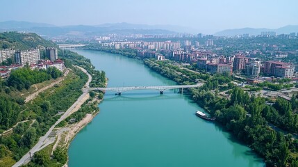 Fototapeta premium High-angle view of a river flowing through a city, with a bridge spanning it. Lush green hills and buildings surround the river
