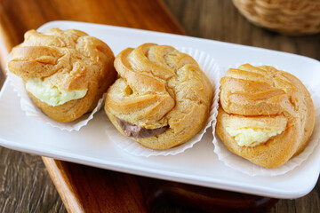 Three freshly baked Kue Sus (Choux Pastries or Cream Puffs) neatly arranged on a  white rectangular plate. Each puff is perfectly golden-brown and filled with a visible, creamy filling.