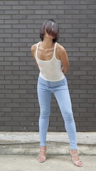 A young Asian female model in blue skinny jeans and a white tank top stands against a textured...