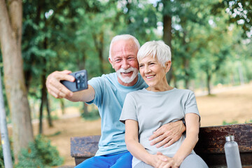 Happy active senior couple having fun using smartphone, taking selfie, or having a video call and wearing sportswear, after having an exercise sport activity outdoors