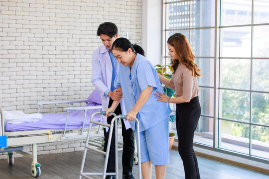 Asian man physiotherapy doctor help support senior woman patient injury practice using walker frame to walk slowly in hospital with lovely daughter, caregiver specialist wellness healthcare treatment.