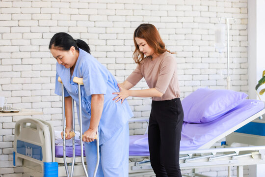 Asian young woman teaching walking support crutches walks beside senior female patient in hospital bed room medical recovery caregiver specialist wellness healthcare treatment rehabilitation support.