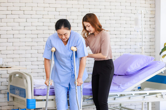 Asian young woman teaching walking support crutches walks beside senior female patient in hospital bed room medical recovery caregiver specialist wellness healthcare treatment rehabilitation support. - Powered by Adobe