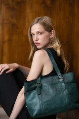 A fashion model with long wavy blonde hair is captured in a full-length, grounded pose, resting against a textured wood-paneled wall while showcasing a dark teal leather tote bag