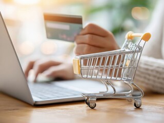 A person holds a credit card over a laptop keyboard, surrounded by soft lighting. A small shopping cart is positioned in the foreground, indicating online shopping activity.