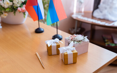A wooden table displays two gift boxes with ribbons, a decorative box, and two flags. The setting is bright and inviting, suitable for a celebration or event.