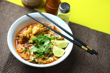 The image shows a bowl of Khao Soi, a traditional Thai noodle curry with chicken, crispy noodles, fresh cilantro, lime wedges, and pickled greens. The dish is beautifully arranged and vibrant.