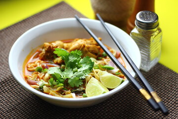 The image shows a bowl of Khao Soi, a traditional Thai noodle curry with chicken, crispy noodles, fresh cilantro, lime wedges, and pickled greens. The dish is beautifully arranged and vibrant.