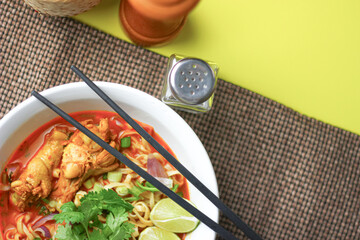The image shows a bowl of Khao Soi, a traditional Thai noodle curry with chicken, crispy noodles, fresh cilantro, lime wedges, and pickled greens. The dish is beautifully arranged and vibrant.