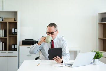 Senior businessman in formal attire using tablet and drinking coffee at modern office desk, working...