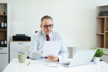 Senior businessman in formal attire using tablet and drinking coffee at modern office desk, working...