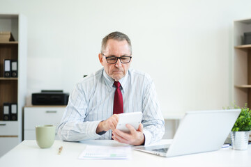 Focused senior businessman holding a document and using calculator at modern office desk, analyzing finance, budget, and corporate planning data with coffee beside.