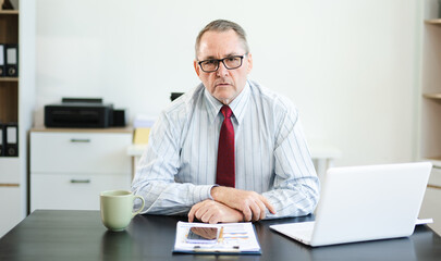 Frustrated senior businessman with smartphone gestures in confusion at office desk, reacting to bad...
