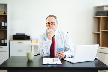 Obraz premium Frustrated senior businessman with smartphone gestures in confusion at office desk, reacting to bad news, tech issue, or business problem with laptop and reports.