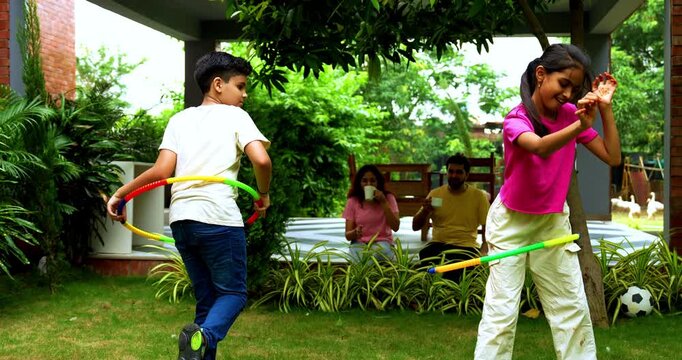 Indian siblings playing hula hoop in home garden while parents laugh and enjoy cheerful family moment, spending quality outdoor time together with fun activities, games, and bonding