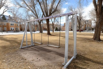 Fototapeta premium Empty wooden swing set dominates quiet urban park in winter. Bare trees, historical residential buildings, clear sky frame scene, evoking feelings nostalgia solitude in forgotten playground area.