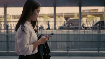 A confident Asian businesswoman walks while using her smartphone in an urban environment, showcasing modern technology, professional success, and digital connection in daily work life. - Powered by Adobe
