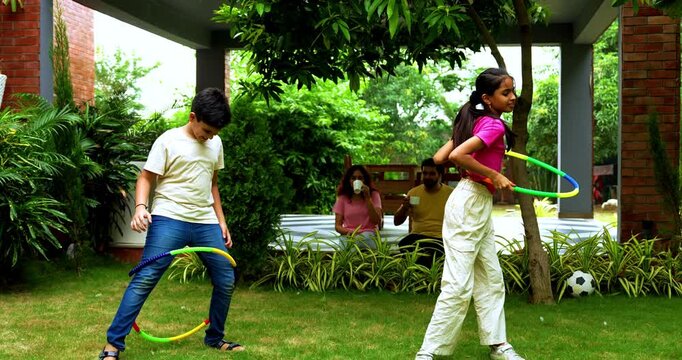 Indian siblings playing hula hoop in home garden while parents laugh and enjoy cheerful family moment, spending quality outdoor time together with fun activities, games, and bonding