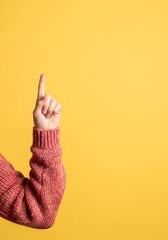 Young adult's hand in a warm sweater pointing upward on yellow background.

