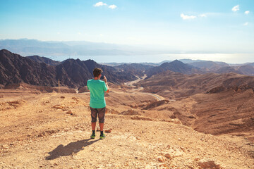 A man travels through the mountain desert. The man takes a photo of a beautiful desert landscape with his mobile phone. Eilat Mountains Reserve in the Negev Desert, Israel