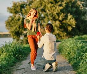 Cheerful handsome man proposing to his girlfriend. Portrait of a young happy couple having fun outdoors in nature during sunset. Girlfriend and boyfriend bonding, love concept during summer.
