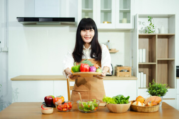 Asian woman shows fresh vegetables with a smile in a modern kitchen. A clean, healthy, and colorful concept perfect for food, wellness, and lifestyle themes.