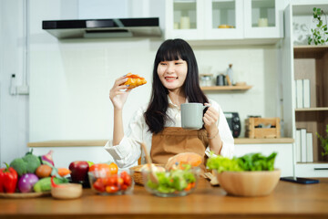 Smiling Asian woman enjoys a croissant and coffee in a modern kitchen with fresh vegetables. A perfect breakfast and lifestyle scene for wellness, food, and home themes.