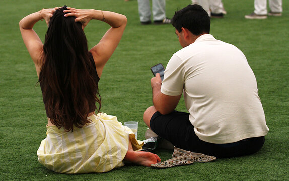 Pareja sentada tranquilamente en la hierba con mujer descalza toc&aacute;ndose el pelo y hombre mirando el tel&eacute;fono m&oacute;vil