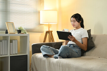 Happy asian woman drinking coffee holding tablet in hand sitting on comfortable couch at home.