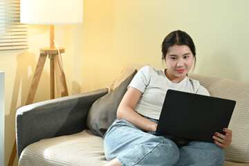 A woman is sitting on a couch with a laptop in front of her. She is wearing a white shirt and blue...