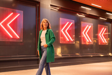 Confident woman in green coat walking past neon arrows at night urban street