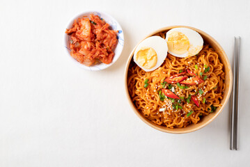 Top view of spicy Korean dried instant noodles with boiled egg, sliced scallions, sesame seed and chili in kraft paper bowl and kimchi cabbage on white background