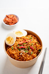 Spicy Korean dried instant noodles with boiled egg, sliced scallions, sesame seed and chili in kraft paper bowl and kimchi cabbage on white background