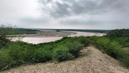 Wide meandering Sawan River in Punjab Pakistan with reddish brown flooding water, sandbanks, vegetated island and dense green banks under hazy overcast sky