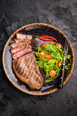 Grilled striploin steak, beef meat steak on a plate with salad. black background. top view