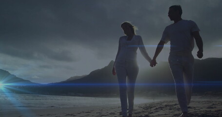 Fototapeta premium Walking mature couple holding hands along sandy shoreline at dusk, with dramatic sky and lens flare