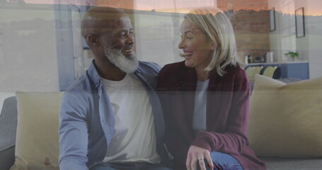 Smiling senior couple sitting closely on sofa in living room, with large window sunset view
