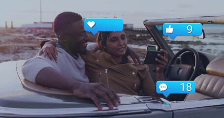 Couple embracing while checking smartphone in convertible on coast at dusk, with floating icons