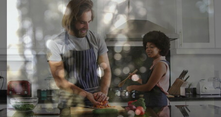 Man wearing apron slicing carrots and woman stirring pot with wooden spoon in kitchen with greens