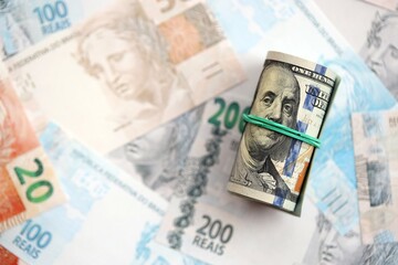 Roll of US dollar bills lies on big amount of Brazilian reais banknotes close up