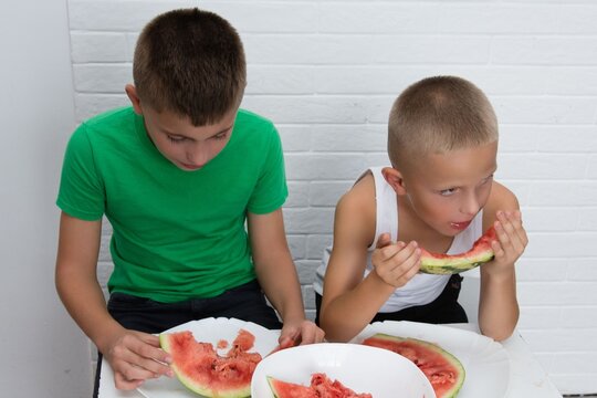 Two boys are enjoying watermelon. The siblings are sitting in a room with plates and eating the refreshing summer fruit, with a white brick background. - Powered by Adobe
