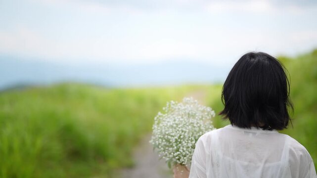 Japanese woman in a white dress walks away in a summer grassland under a clear blue sky, with a gentle breeze ruffling her hair, holding a bouquet of baby's breath. Nagano Prefecture, Japan. Summer.