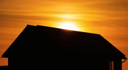 silhouette of a house with solar panels against an orange sunset sky