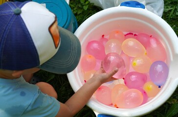  Child holding a colorful water balloon, preparing for a playful summer game. A bucket filled with balloons ready for excitement and splashes on a green outdoor lawn.