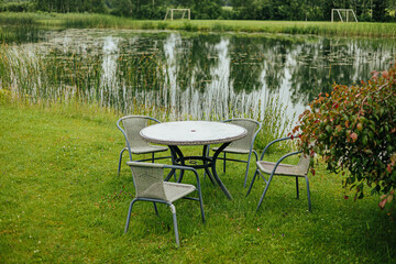 Empty outdoor tables and chairs arranged on a green lawn by a calm lake, surrounded by nature,...