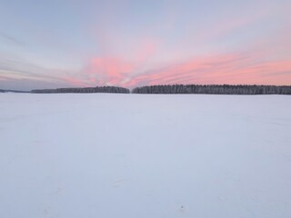Snowy landscape at sunset featuring a vast field and distant trees under a pink sky glow